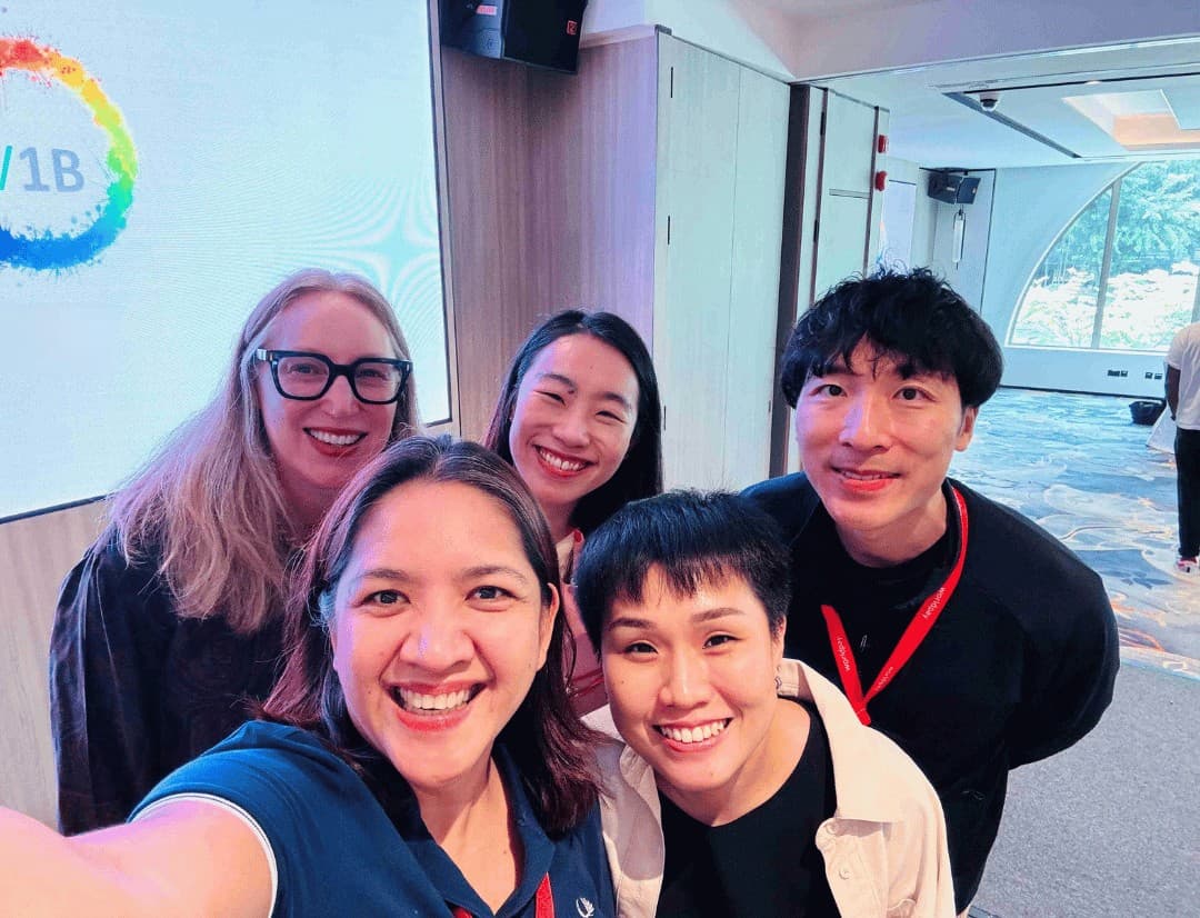 Four women and one man taking a selfie smile towards the camera in an office. A rainbow circle with ‘V1B’ in the middle is projected behind them on a whiteboard.