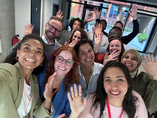 a diverse group of colleagues waving at the camera in a group selfie in an office building