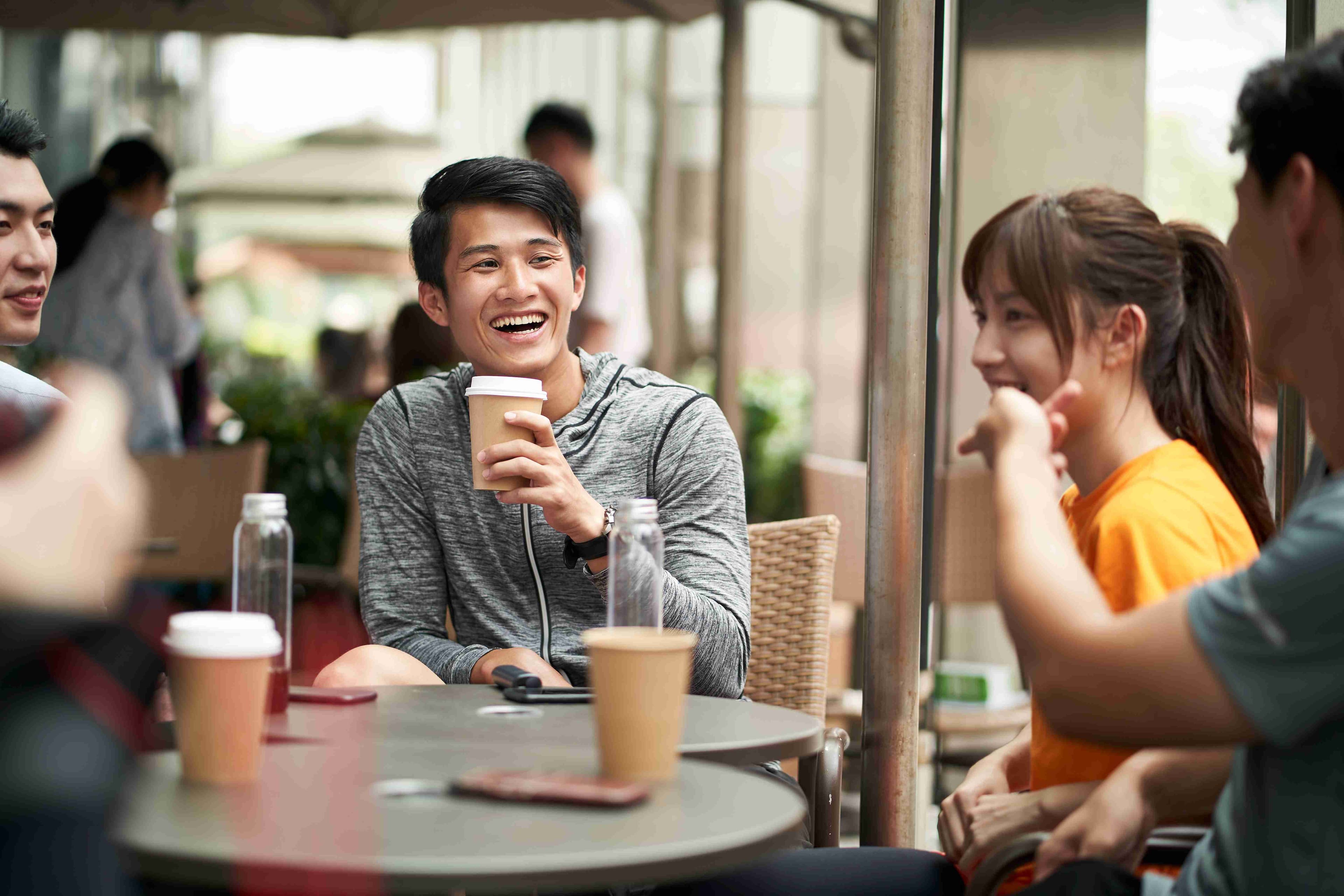 Group of colleagues chatting and laughing around an outdoor café table with takeaway coffees.