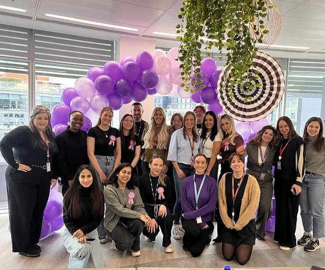Group of people posing in an office with purple and white balloon arch, under hanging plants. A Worldpay banner with company values is visible on the right.