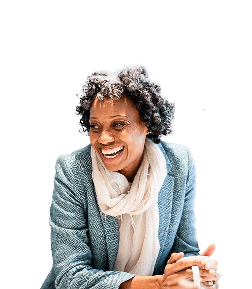A woman leans on a table in a green blazer and white scarf and smiles at something off camera. There’s a man in a blue shirt sitting behind her also smiling.